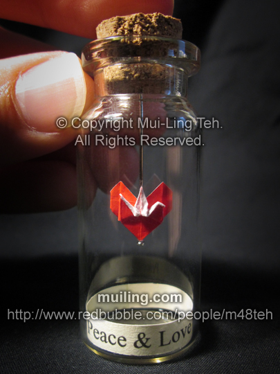 Miniture red origami crane and heart in a bottle by Mui-Ling Teh, with the message "Peace & Love" written below on a yellow label. The crane and the heart are made out of one paper.